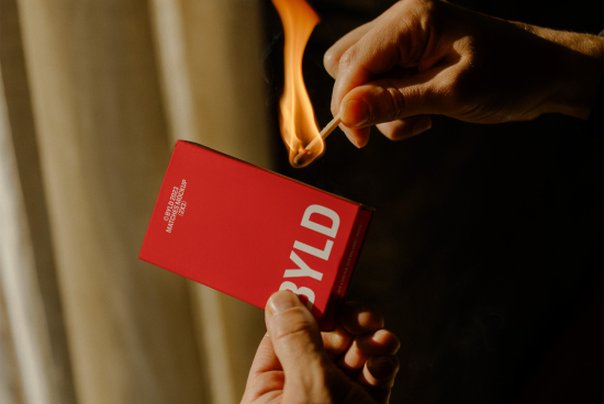Person holding a striking match with dynamic flame near a red matchbox mockup with bold font, ideal for branding and packaging designs.