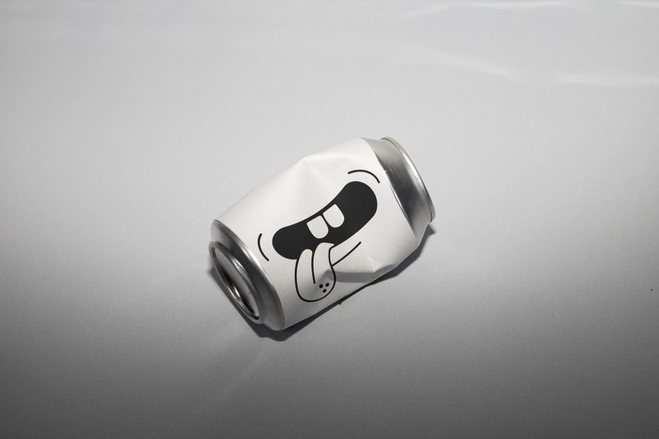 Crushed aluminum can with cartoon face design, lying on a gray surface, realistic mockup for packaging, branding, and advertising.