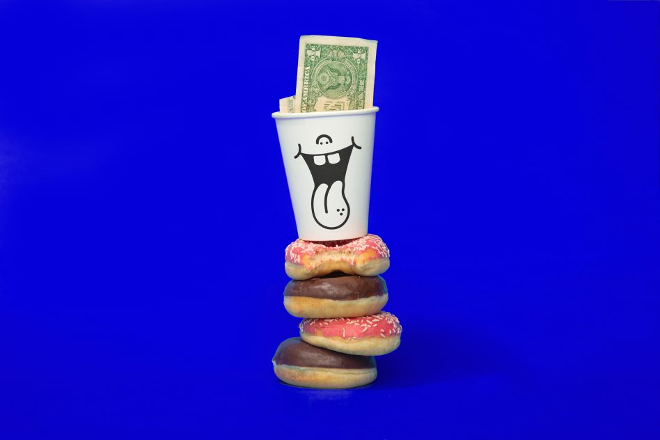 Creative paper cup with happy face design and dollar bill on top of colorful doughnuts, vivid blue background, ideal for mockup graphics.