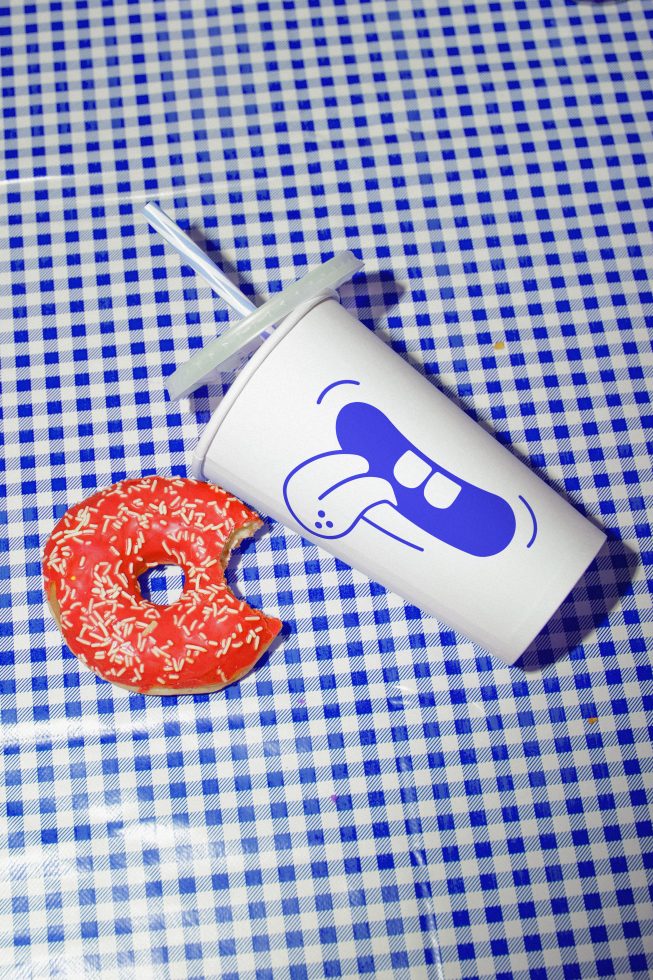 Disposable cup with straw and bitten donut on blue checkered background, ideal for mockup, branding, and design presentation.