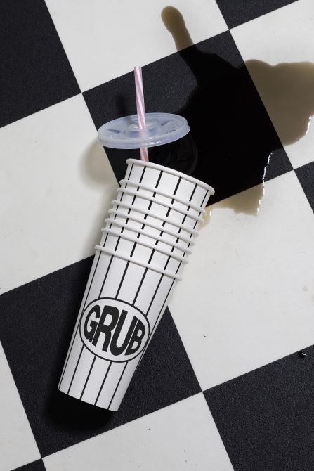 Spilled beverage in a striped paper cup with GRUB logo on checkered surface, ideal for mockup, graphic design, and branding presentation.