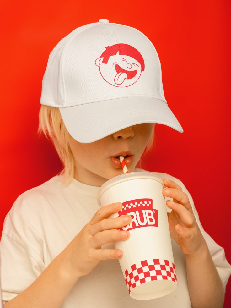 Child wearing white cap with logo drinking from paper cup, red background - creative mockup for branding and advertising designs.