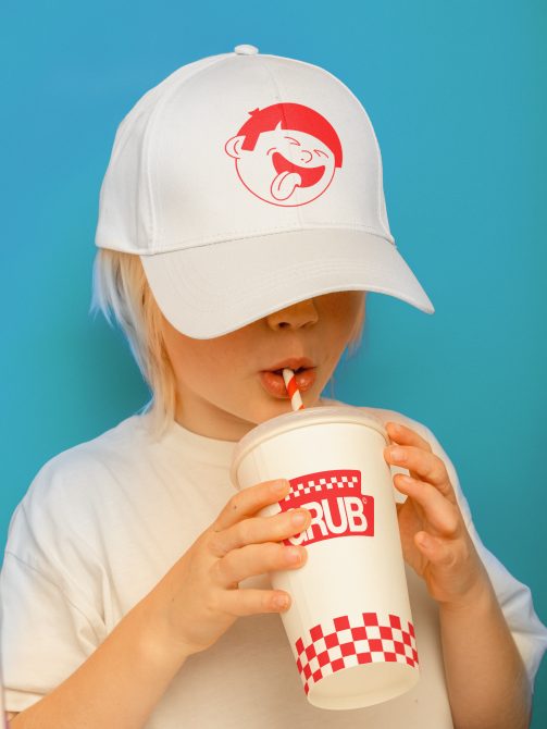 Child in white cap with red logo mockup sipping drink from branded cup, ideal for apparel and branding designs.