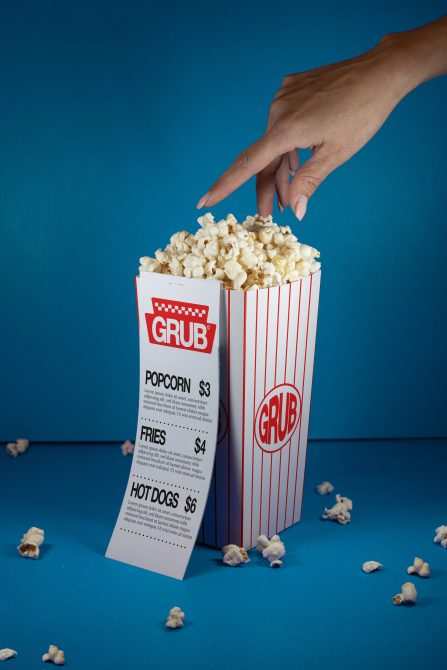 Hand reaching into a striped popcorn box with scattered kernels on a blue background, ideal for mockup and food-themed design graphics.