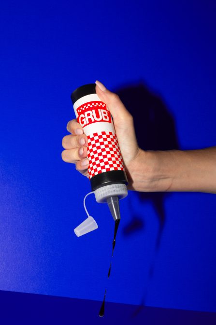 Hand squeezing a bottle with graphic label design mockup, liquid drops falling, on blue background, ideal for product packaging and branding.