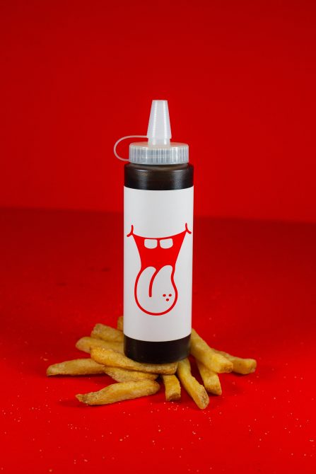 Ketchup bottle mockup with graphic design on red background, surrounded by fries, ideal for food packaging and branding presentations.