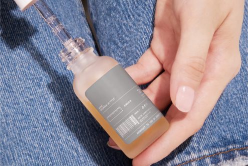 Hand holding cosmetic bottle with label design mockup, product packaging template, skincare beauty branding, close-up denim background.