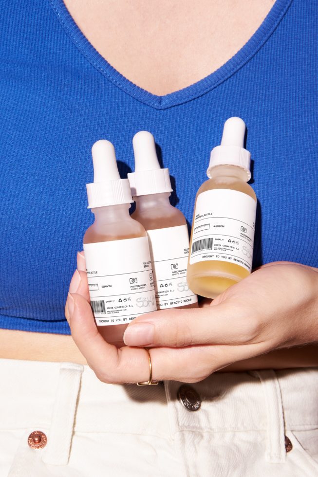 Woman holding three skincare serum bottles, cosmetic mockup, packaging design, with white cap, clear labels, on blue top and white pants background.