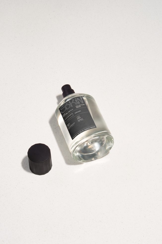 Mockup of a clear glass cosmetic bottle with black label and cap, on a light textured background, perfect for branding presentations.