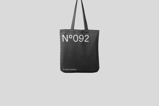 Black tote bag mockup with white text design, hanging against a gray background, ideal for product presentation and graphic design.