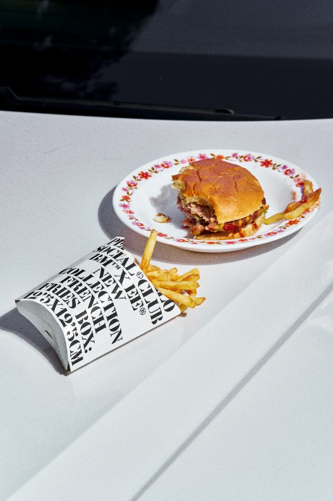 Half-eaten burger on decorative plate with fries spilling from paper container, all atop a white car surface, under sunlight for graphics design.