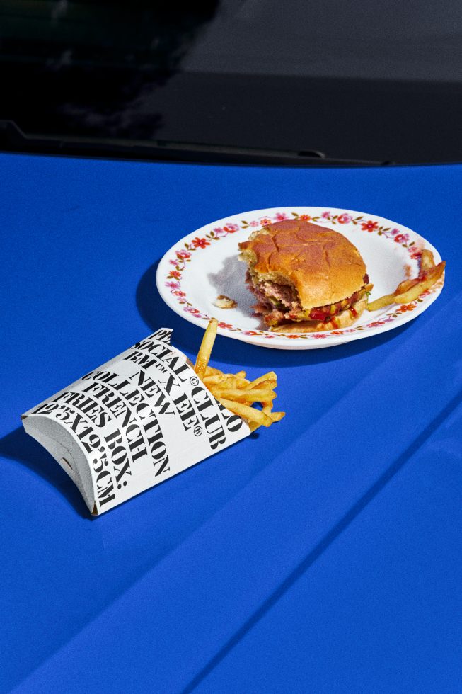 Half-eaten burger on decorative plate with spilled fries and typography newsprint, vibrant blue backdrop, ideal for mockup, food graphics, ad templates.