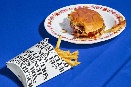 Partial burger on floral plate with spilled fries from paper container on blue background, ideal for mockup and graphic design assets.