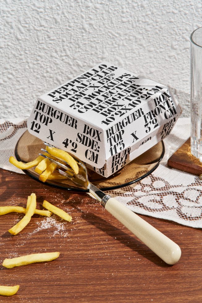 Stylish burger packaging mockup with typographic design on plate, scattered fries and fork, ideal for food-related graphic presentations.