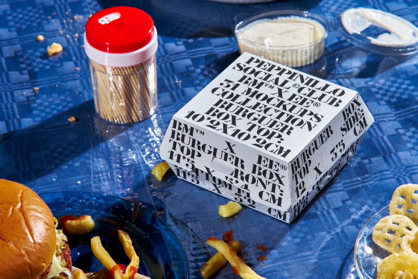 Creative burger packaging mockup with bold font design next to fast food items on a textured blue tablecloth, ideal for graphic designers.