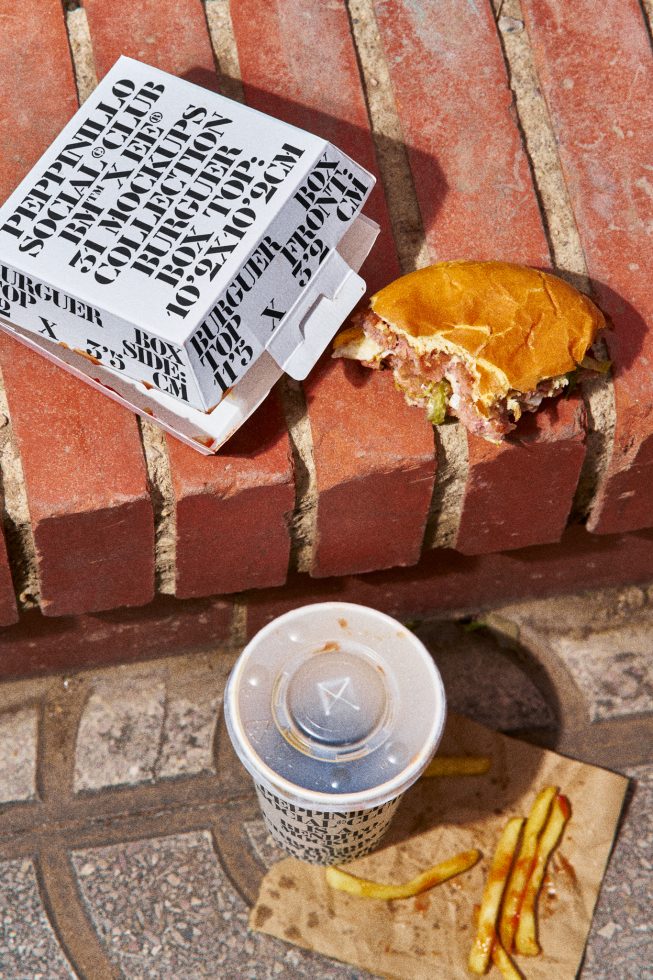 Fast food packaging mockup with typography design on a sunny brick surface, half-eaten burger and drink for product display.