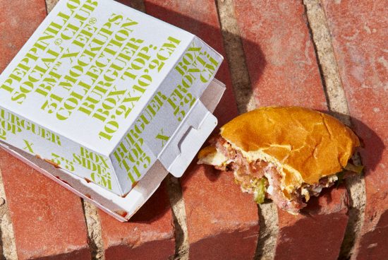 Stylish packaging design mockup featuring a modern typographic burger box next to a half-eaten burger on a brick surface, perfect for presenting brand designs.