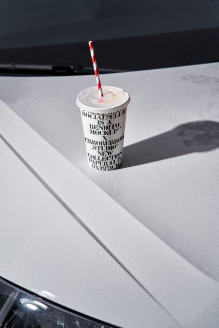 Paper cup with straw on a car hood under sunlight creating shadows, ideal for packaging mockup design, realistic product display.