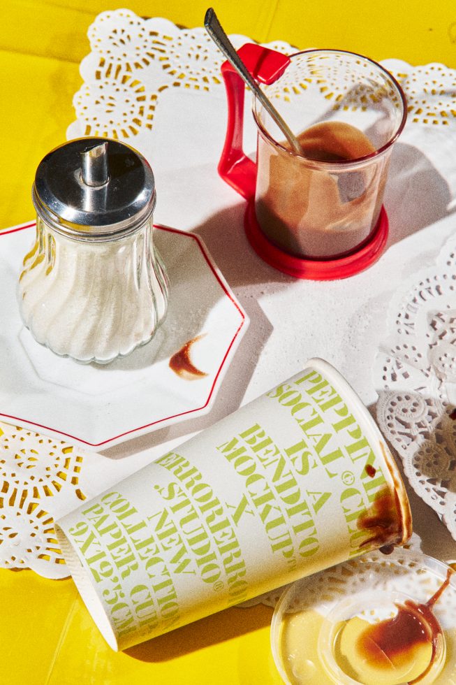 Sunlit moody mockup with spilled coffee, sugar dispenser, and retro glass, featuring bold typography on a paper cup, ideal for graphic design showcase.