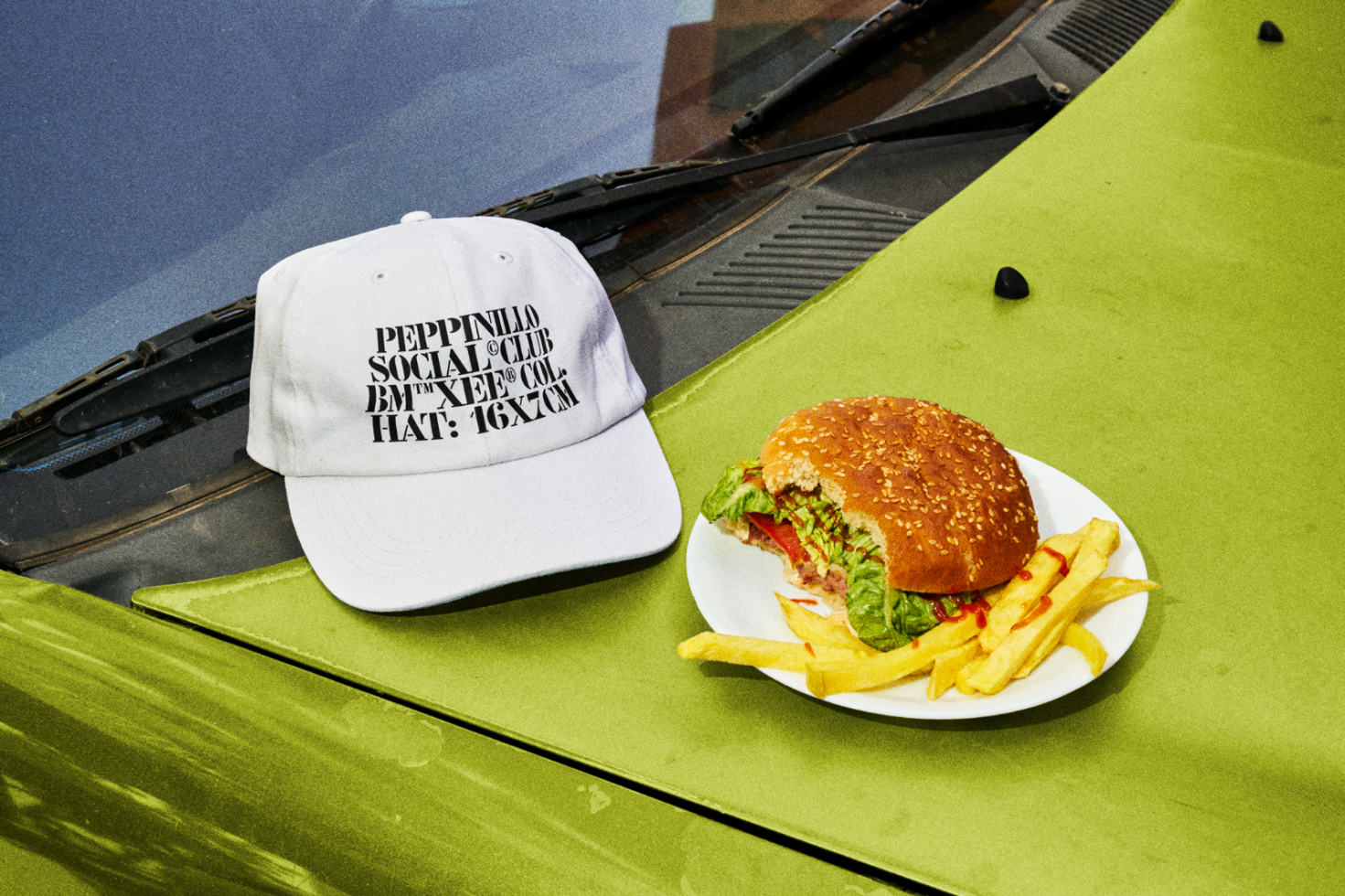 Urban streetwear cap mockup on a car hood with a burger and fries, ideal for showcasing apparel designs in a casual, modern setting.