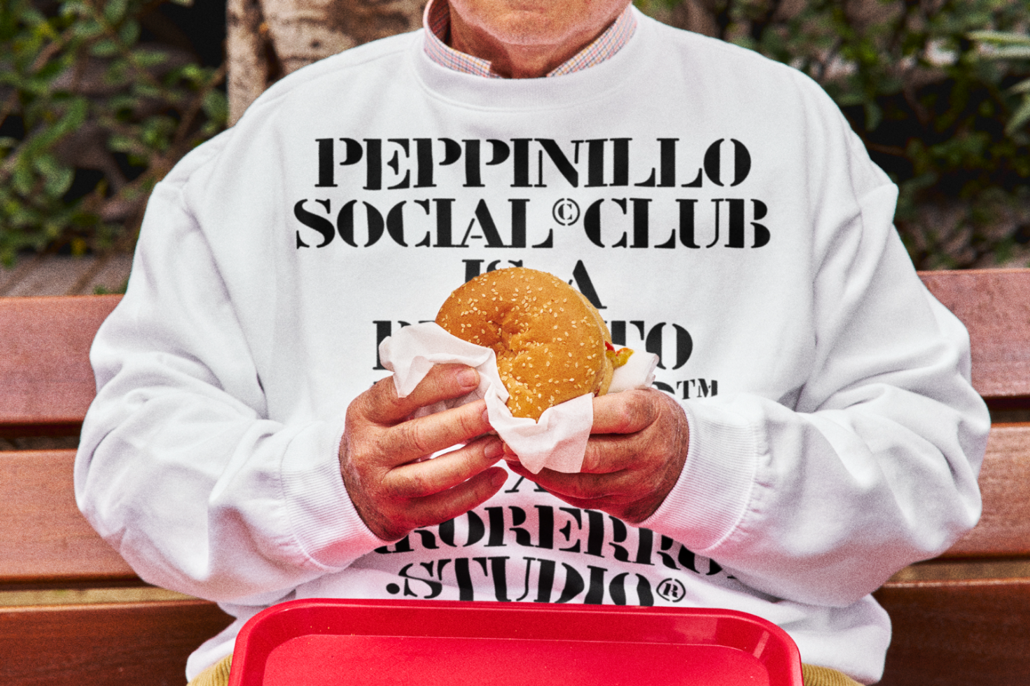 Person in a white branded sweatshirt sitting on a bench holding a burger, streetwear design mockup for branded apparel.