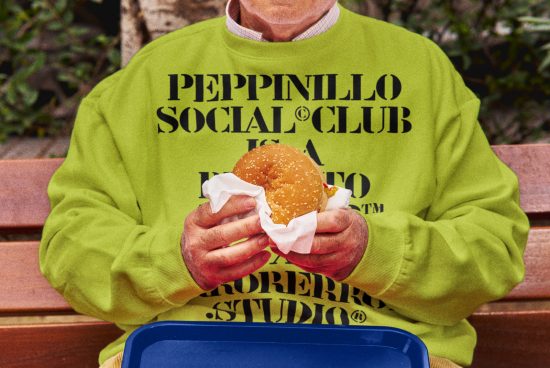 Person in neon green sweatshirt with bold typography design holding a sandwich, sitting on bench, ideal for mockup, graphics, and apparel design.