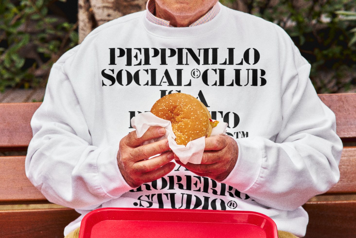 Person in branded sweatshirt holding burger, sitting on bench, detailed texture, realistic mockup for apparel design presentation.