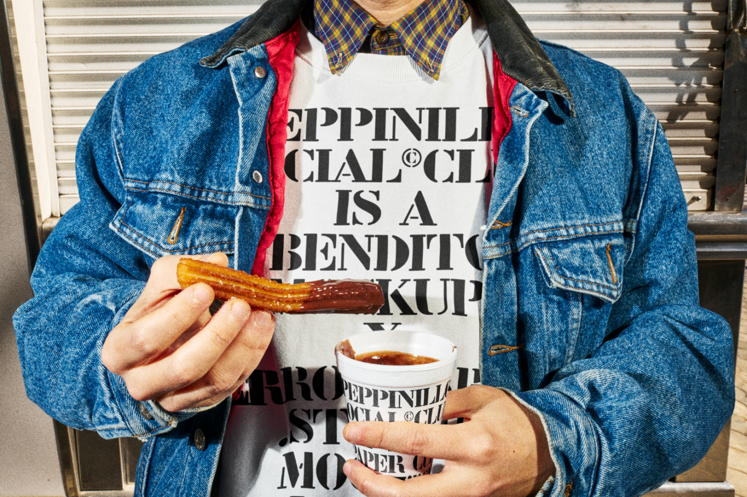 Person in denim jacket holding chocolate-covered churro, urban background, creative streetwear mockup design, trendy fashion graphic.