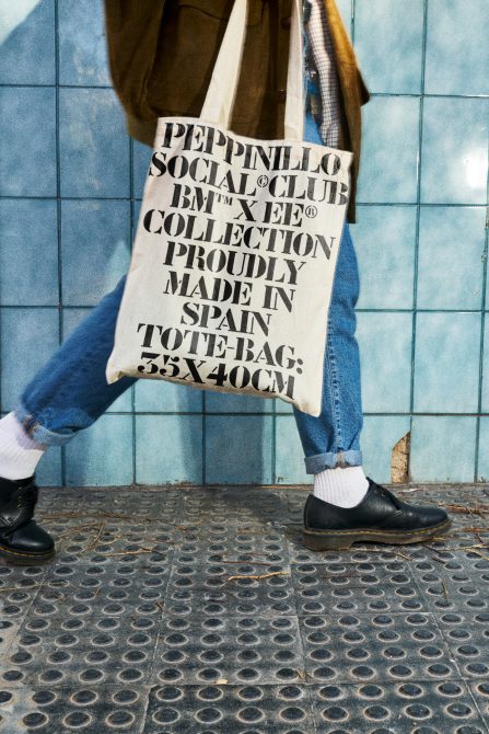 Stylish tote bag mockup with bold typography design, held by person in casual outfit against blue tiled wall, highlighting urban street style.