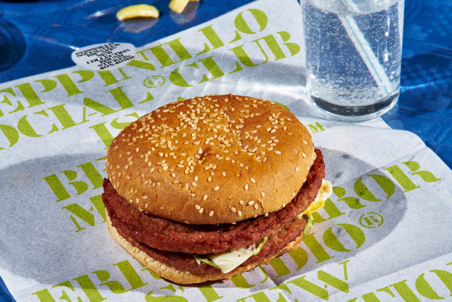 Realistic food mockup with a textured double veggie burger on branded paper, clear water glass, and fries, ideal for restaurant menu design.