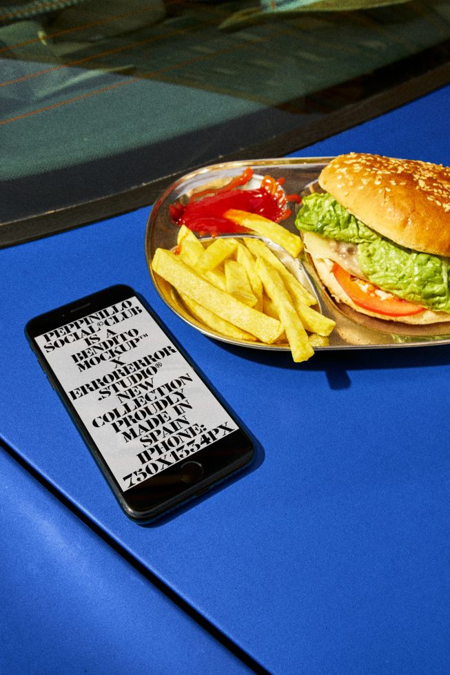 Smartphone on blue surface displaying stylish font, next to burger and fries, ideal for mockups, typography, and food-themed design projects.