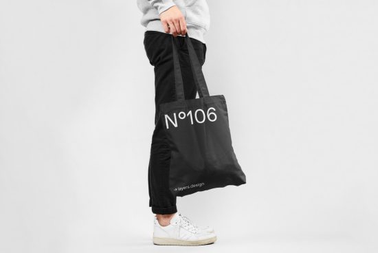 Person holding black tote bag mockup with text and logo, plain background, product design template for ecommerce.