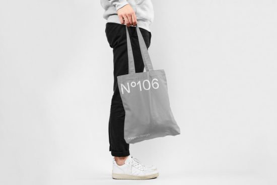 Person holding tote bag mockup with number design on gray background, ideal for showcasing branding designs and patterns.