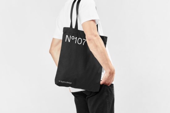 Side view of a person in white t-shirt holding black tote bag with white text mockup, plain backdrop, ideal for designers graphics display.