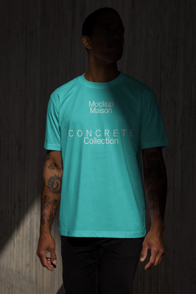 Man in teal t-shirt for apparel design mockup, dramatic lighting, with tattooed arms, against textured concrete backdrop for graphic designers.