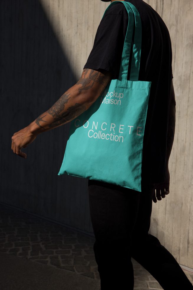 Man wearing black with teal tote bag mockup walking in sunlight, showcasing urban street fashion, ideal for designer graphics.