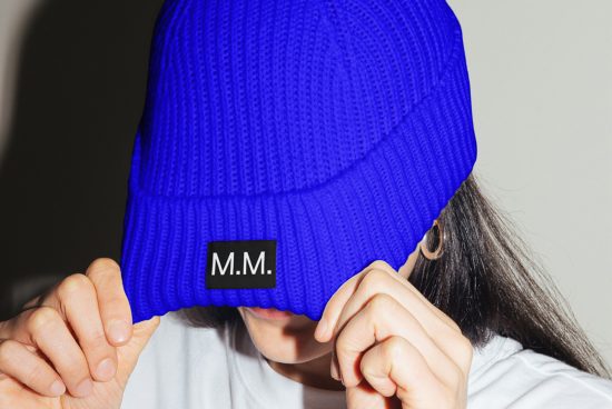 Person pulling a blue beanie over eyes, showcasing a bold label with initials M.M. Ideal for mockup graphics related to fashion design.