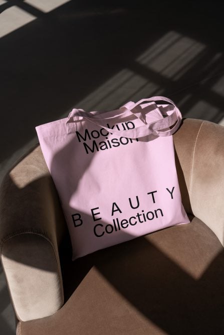 Pink tote bag with bold text Mockup Maison Beauty Collection on beige armchair in sunlight. Ideal for branding mockups tote bag designs digital assets.