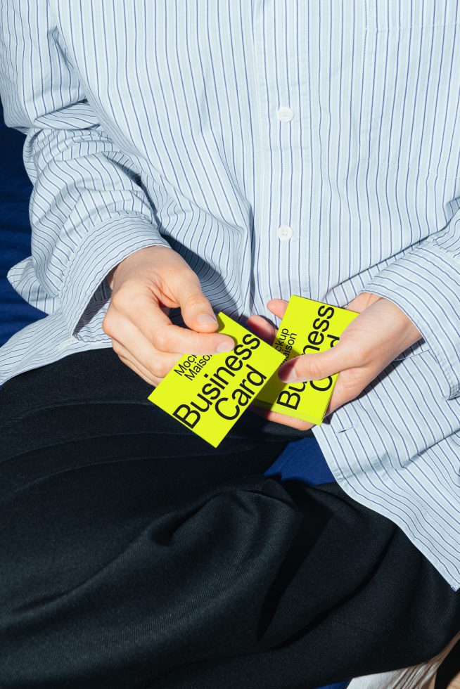 Person in striped shirt holding bright yellow business cards with mockup text for design presentation, close-up on hands and cards against blue background.