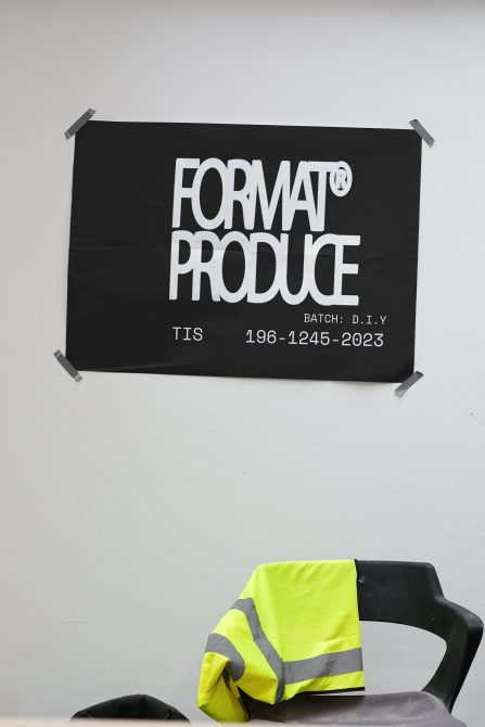 Black poster with FORMAT PRODUCE text taped to wall, yellow safety vest on black chair in front. Ideal for designers needing graphics or typography templates.