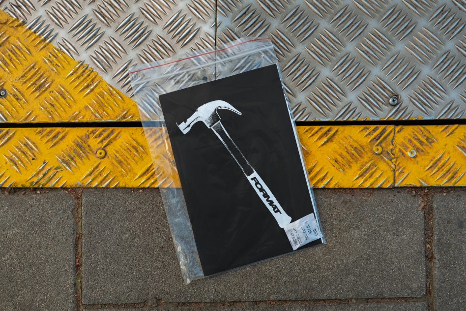 Urban graphic design mockup featuring a hammer illustration on a black background, placed in a transparent bag on a textured metal and concrete surface.