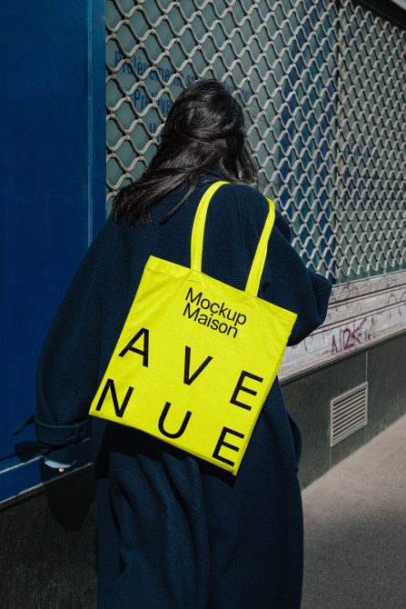 Mockup of a yellow tote bag with black text over a person's shoulder against an urban background. Perfect for designers. Keywords: Mockup, bag, tote, urban.