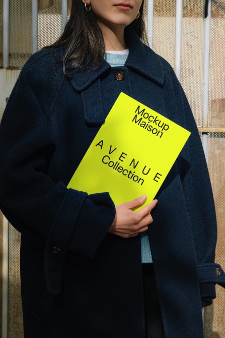 Person holding a yellow magazine with the text Mockup Maison Avenue Collection, wearing a navy coat, suitable for mockup graphic design, poster, marketing.