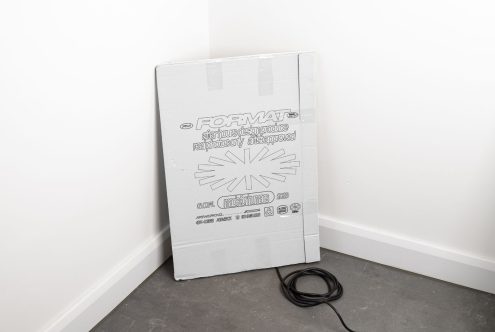 Cardboard packaging for artwork corner protectors leaning against a white wall, black cord on floor. Designers use for mockups product photography studio settings.