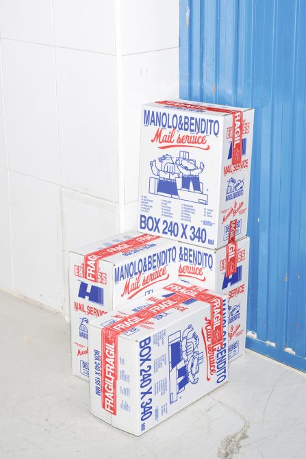 Stacked white cardboard boxes with Manolo&Bendito Mail Service branding, showing in a warehouse; packaging mockup, delivery design, shipping graphics.