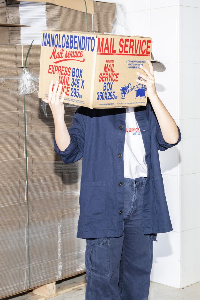 Person holding a large mail service box above their head, standing next to stacked cardboard boxes in a warehouse. Ideal for packaging mockup design.