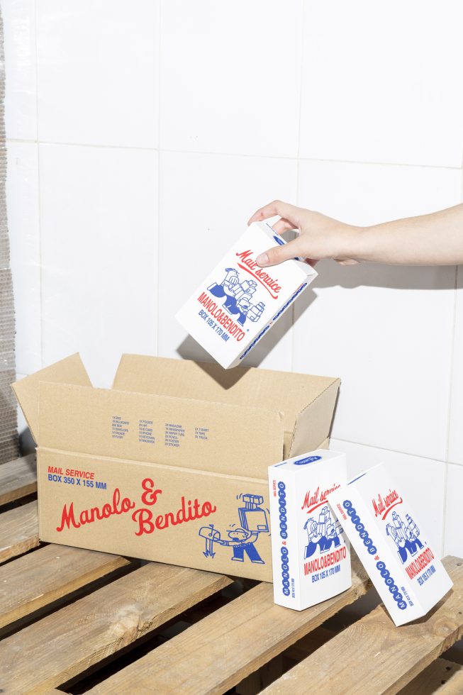 Mockup of cardboard packaging boxes with playful blue red graphics showing Manolo and Bendito branding placed on a wooden pallet, designer asset