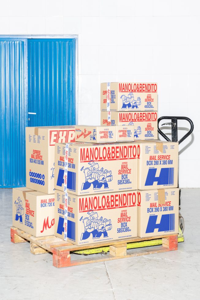 Stacked shipping boxes, labeled Manolo & Bendito Mail Service, on a pallet in front of blue doors in a warehouse setting suitable for mockup designs