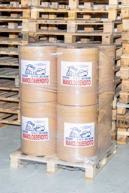 Pallet of large sticker rolls labeled Manolo & Bendito in warehouse setting, suitable for packaging mockups, industrial design templates, packaging graphics.