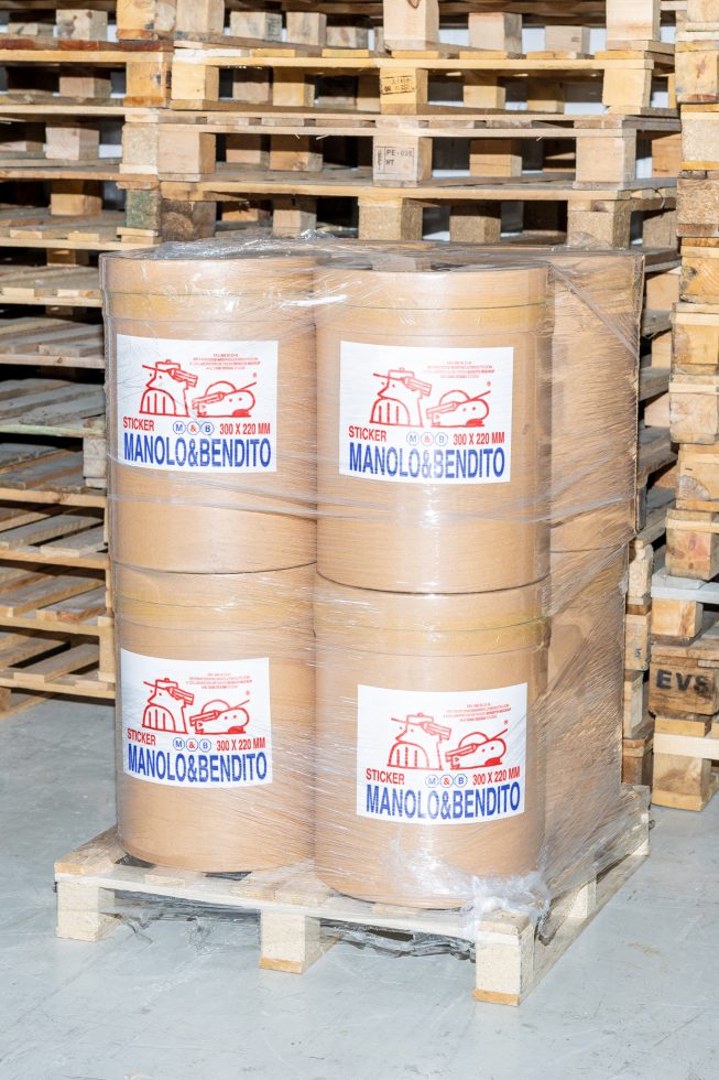 Four large rolls of Manolo and Bendito stickers on a wooden pallet covered in plastic wrap in a warehouse for wrapping and shipping purposes. Mockups, Graphics